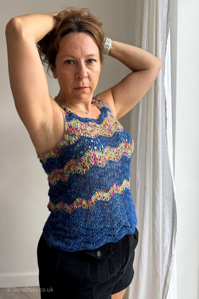 Dora is seen facing the camera turned slightly to the side, holding her hair up to show off the side and underarm views of the light waves crochet vest top pattern. The Pattern is striped with silk yarn in dark blue mixed with a variegated rainbow yarn.