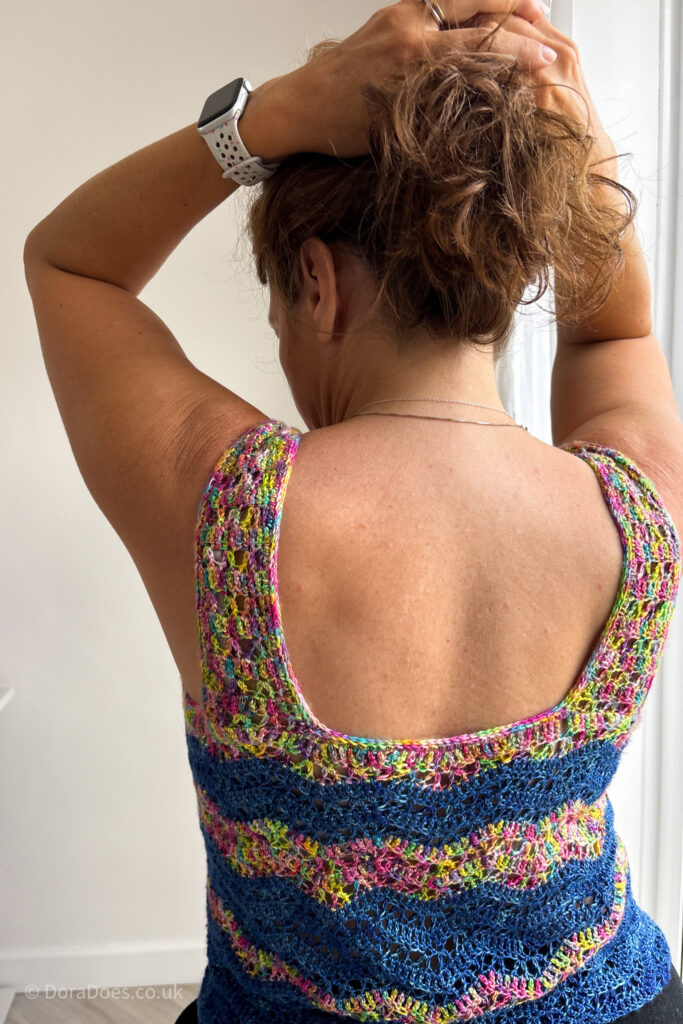 A view from behind of a woman wearing a striped ripple crochet top. She holds her hair up to show the back of the vest