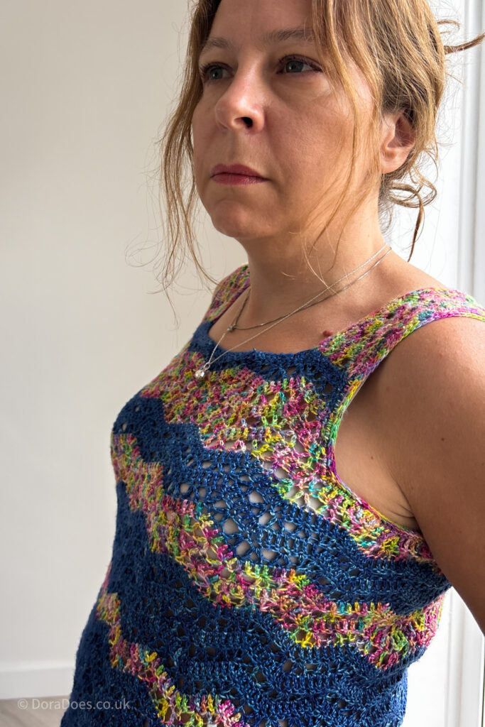 Dora is seen from the side wearing a silk crochet vest top which has a lace chevron stripe stitch pattern in a dark blue yarn paired with a multicoloured rainbow yarn.