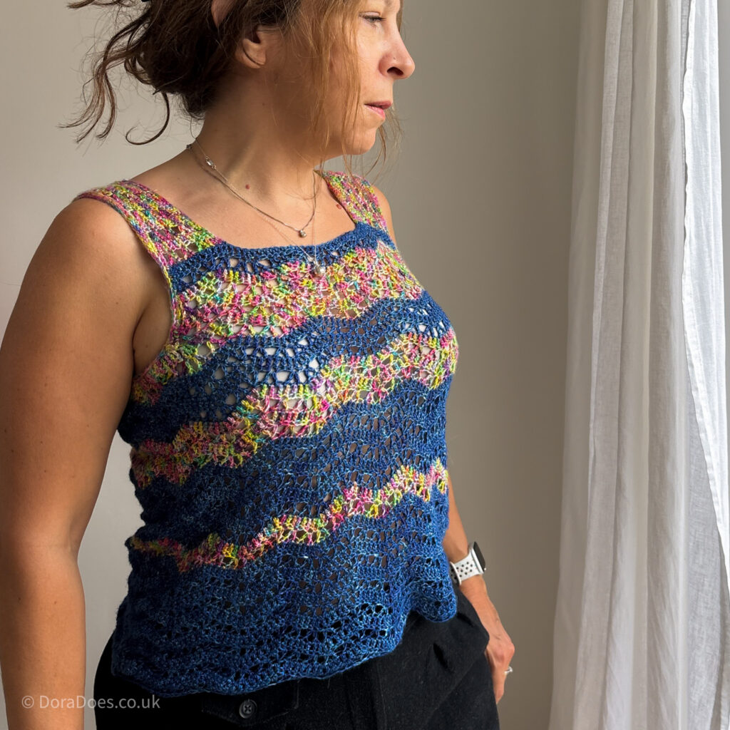 A square image showing Dora wearing a multicoloured silk crochet vest top. The crochet top has asymmetric stripes in a crochet ripple stitch. The Stripes are dark blue mixed with a variegated rainbow yarn.