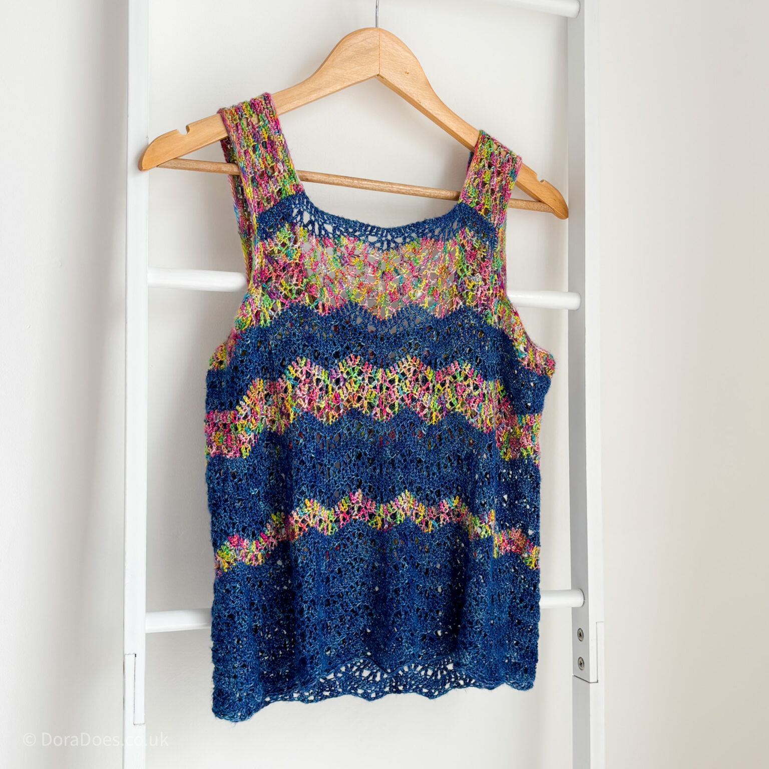 A chevron striped crochet vest top in two colours of silk yarn hangs on a wooden hanger on a blanket ladder in front of a white wall.