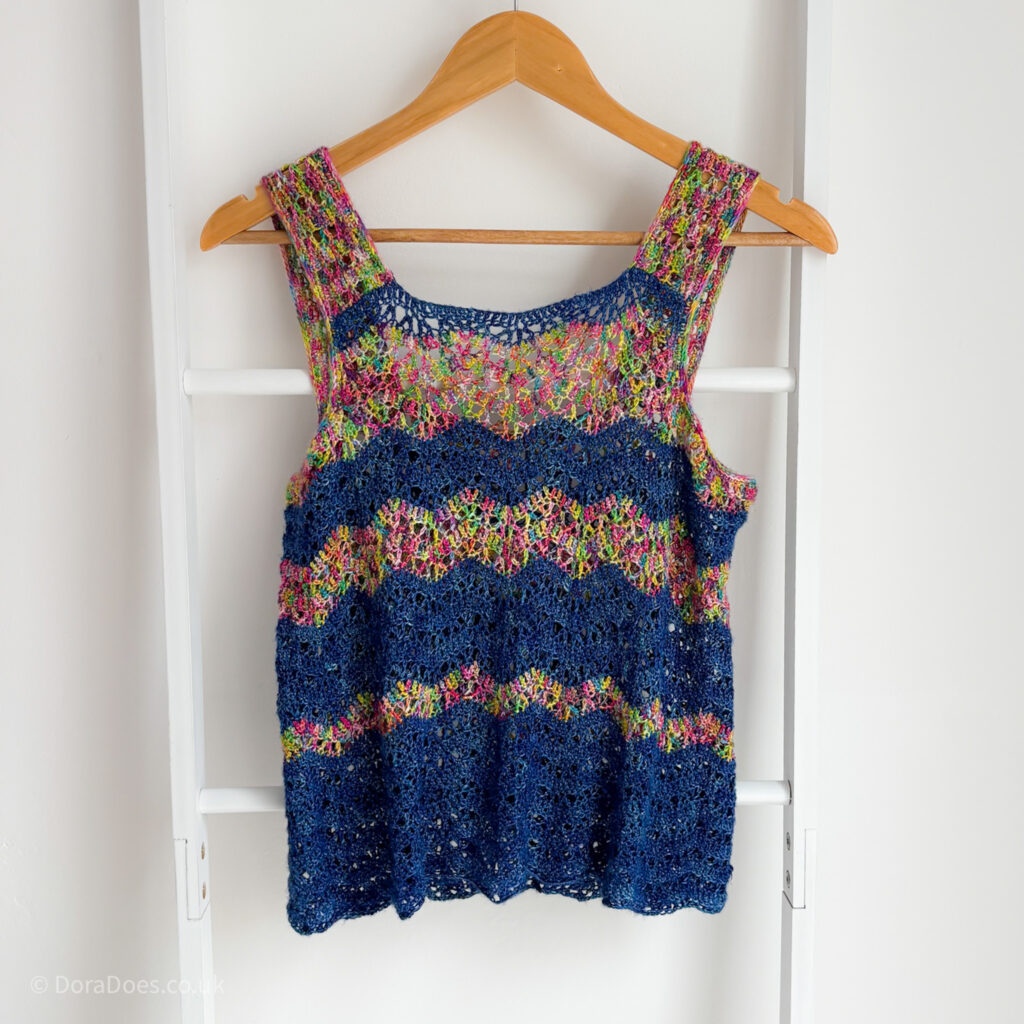 A crochet camisole vest top with thick straps hangs from a blanket ladder on a wooden hanger. The Crochet vest is made from multicoloured rainbow yarn paired with a dark blue. The asymmetric stripes are in a ripple stitch pattern with lace and solid crochet stitches.