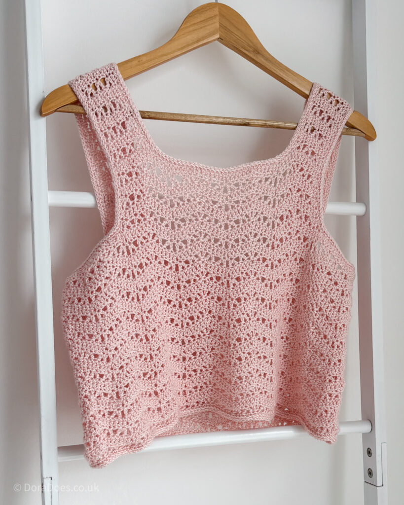 A pink cropped crochet tank top hangs from a blanket ladder. The pink yarn is a blend of wool and silk and is crocheted in a lace wave stitch pattern.