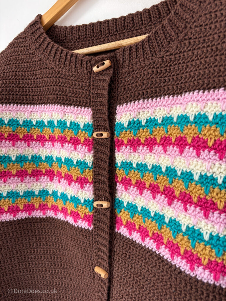 A close up of the stitch pattern on the crime of a brown crochet cardigan with a crew neck and ribbed collar and button band. The interlocking spike crochet stitches form horizontal stripes in shades of pink, teak, cream and mustard yellow. The cardigan has a slightly retro feel about it.