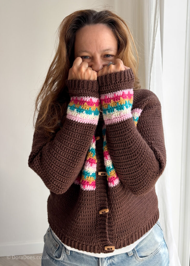 Dora wears a brown crochet cardigan with pink, yellow and teal stripes across the chest and cuffs. She holds the long sleeves up to her fact to show how excited she is by sweater weather.
