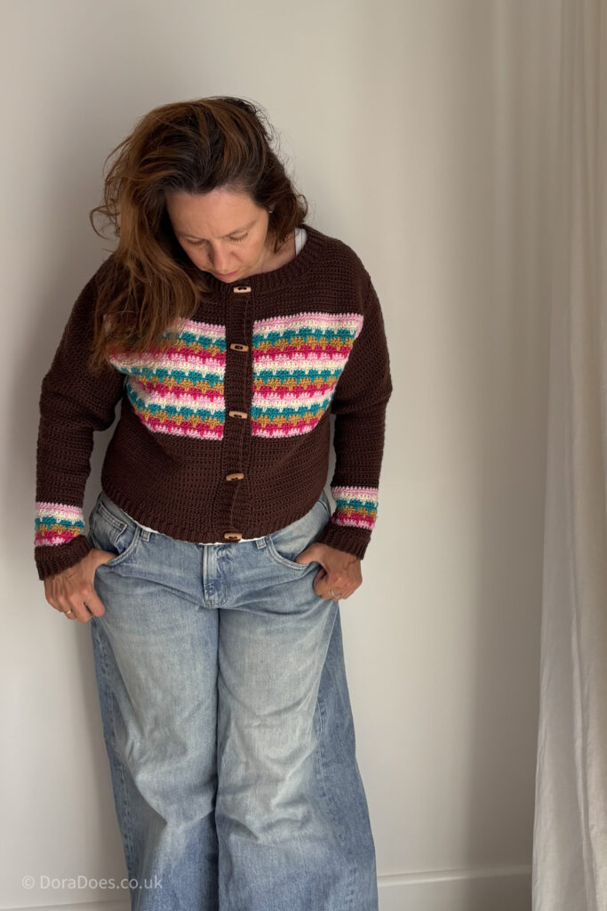 Dora leans against a white wall wearing a crochet cardigan paired with baggy jeans. The relaxed fit cardigan is mostly brown with pink, cream, mustard yellow and turquoise horizontal stripes which interlocking made of larksfoot style spike crochet stitches.