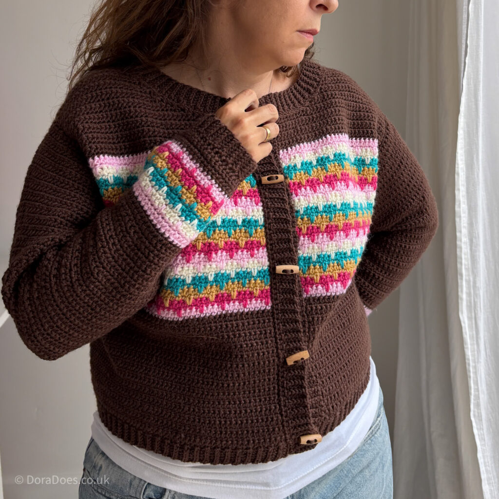 A brown crochet cardigan with colourful horizontal stripes, ribbed cuffs, collar, hem and button band, with toggle buttons is seen close up, modelled by Dora. One hand is on her hip and the other moves to undo the top button.