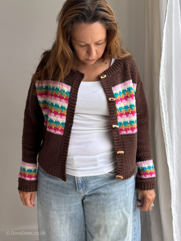 Dora is seen front on wearing a brown crochet cardigan with colourful horizontal stripes across the chest. The cardigan is unbuttoned.