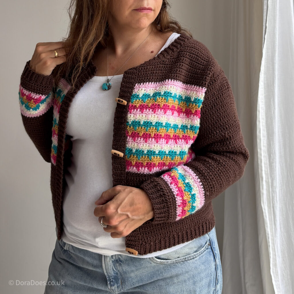 The torso of a woman wearing a crochet cardigan with colourful interlocking stripes across the chest and a matching stripe pattern on the wrists set against a brown background.