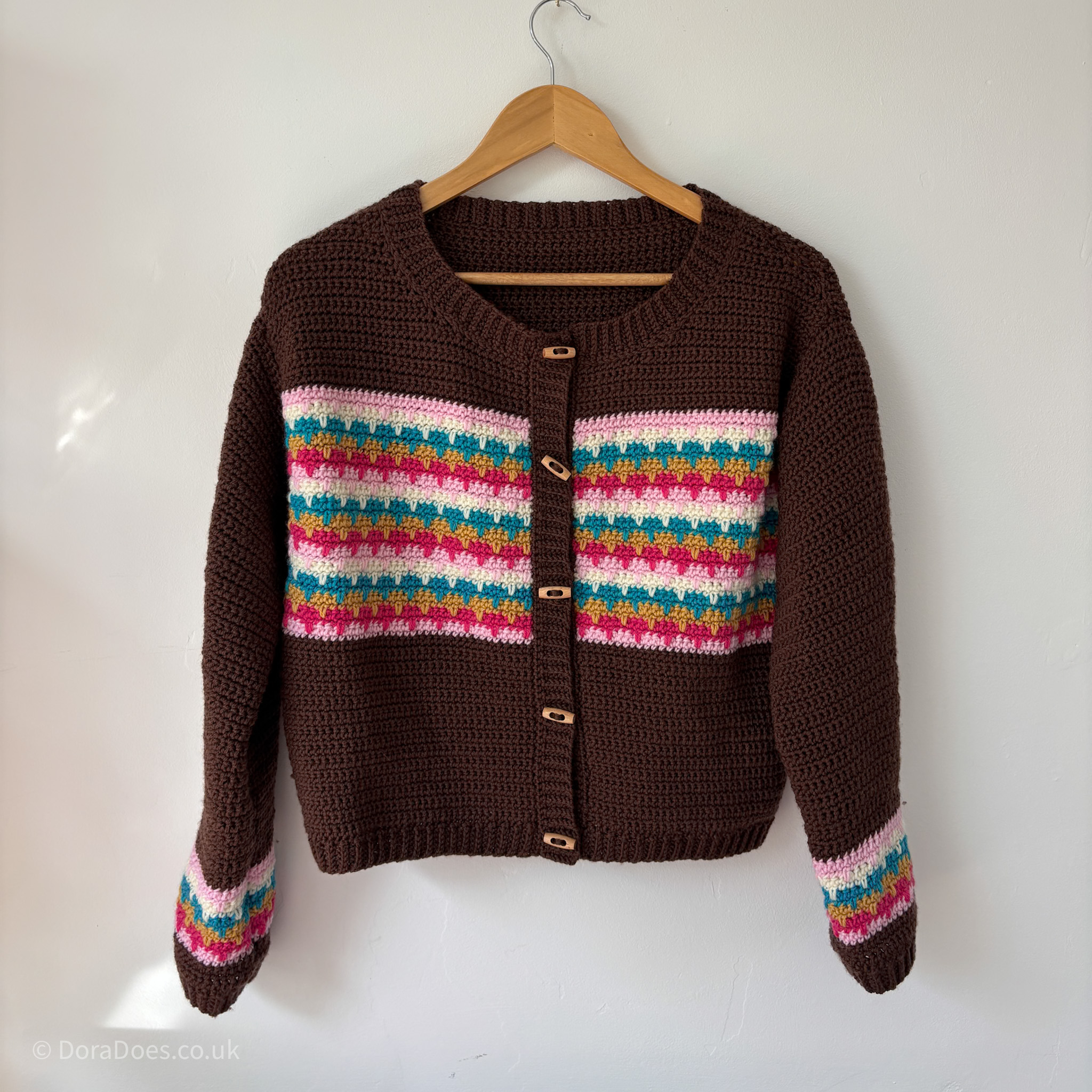 A brown crochet cardigan with colourful stripes of pink, cream, modified and teal across the chest and wrists is hanging from a white wall. The crochet cardigan has long sleeves with ribbed cuffs, ribbing round the hem, collar of a round neck and the button band. The cardigan has 5 wooden duffel style buttons down the front.