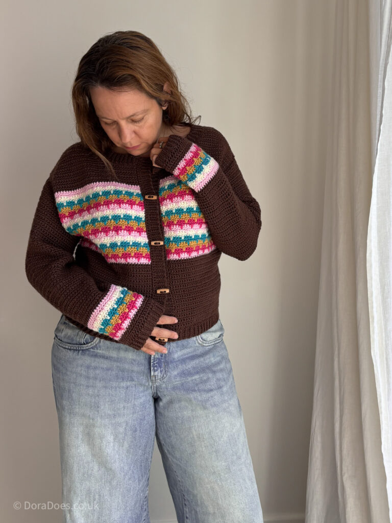 Dora wears a cozy crochet cardigan paired with jeans. The brown cardigan has a bright colourwork pattern of stripes across the chest and matching pattern on the wrists of the long sleeves.