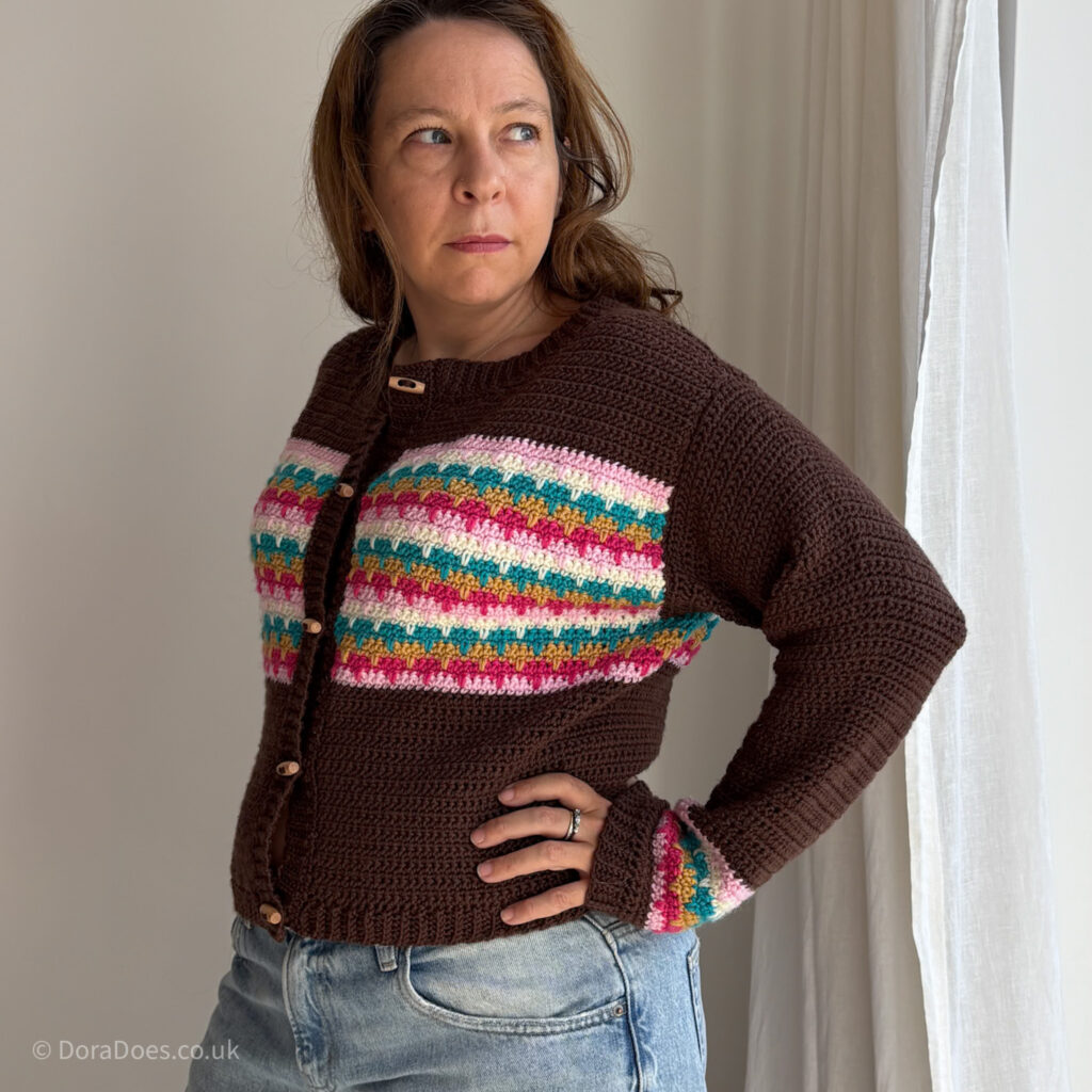 Dora stands side on, one hand on hip, wearing the lark crochet cardigan. The brown cardigan has colourful stripes across the chest and wrists, 5 wooden buttons down the front, long sleeves, a round neckline and ribbed crochet cuffs, hem, collar and button band.