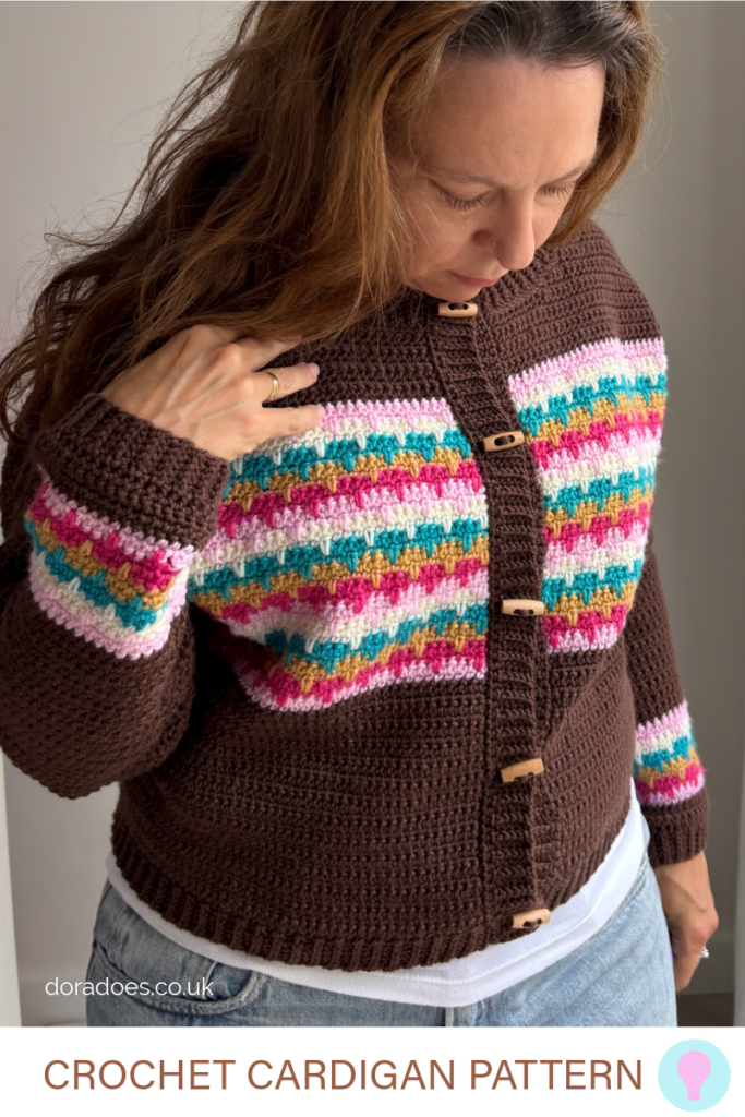 A close up of the front of the crochet ‘lark cardigan’ worn by Dora, who holds one. Hand to her shoulder whilst looking at the wooden duffle buttons on the cardigan. The brown cardigan has colourful stripes around the chest and wrists with simple crochet rib stitches around the cuff, bulge, hem and button band. A text overlay reads Crochet Cardigan Pattern.