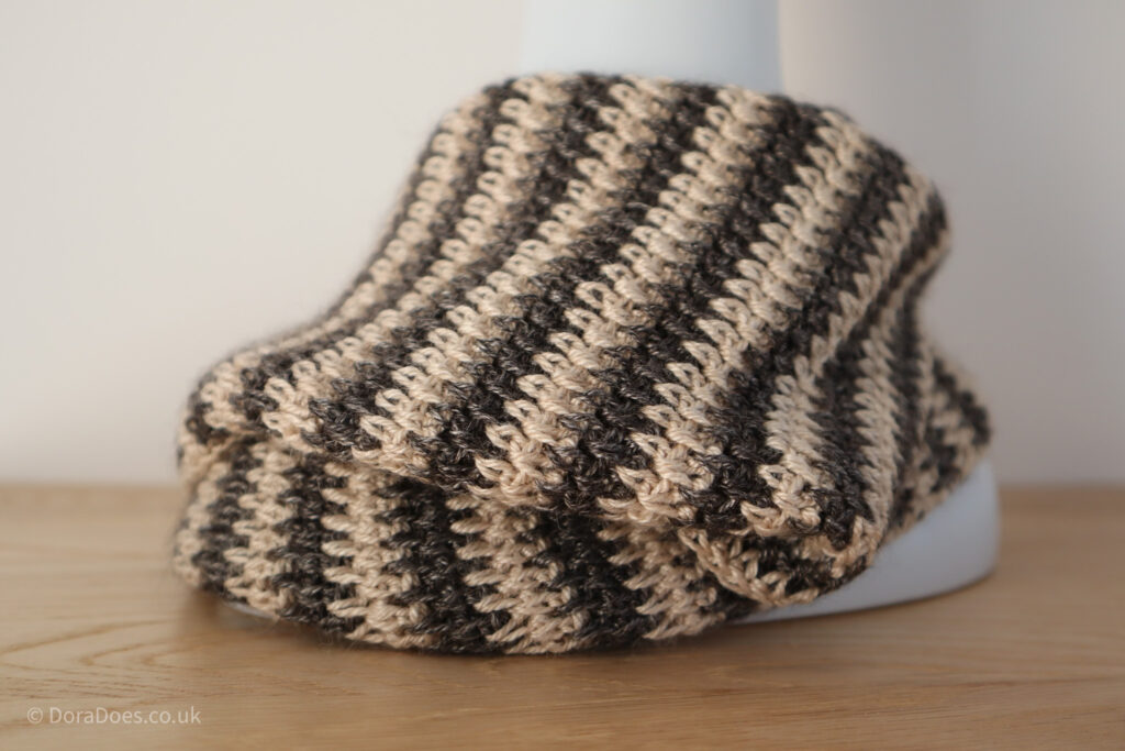 A close up of the stitches in a folded over crochet cowl draped around the base of a mannequin. The stripes are made in pewter and cream yarn with the stitches interlocking between the posts.