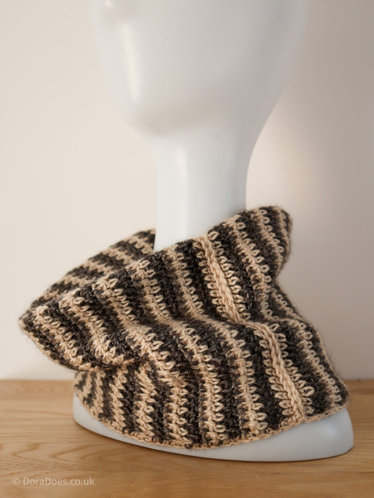 A portrait image of a white head and shoulder mannequin adorned with a brown and natural cream stripy crochet cowl which is worn doubled over with a visible seam along one side.