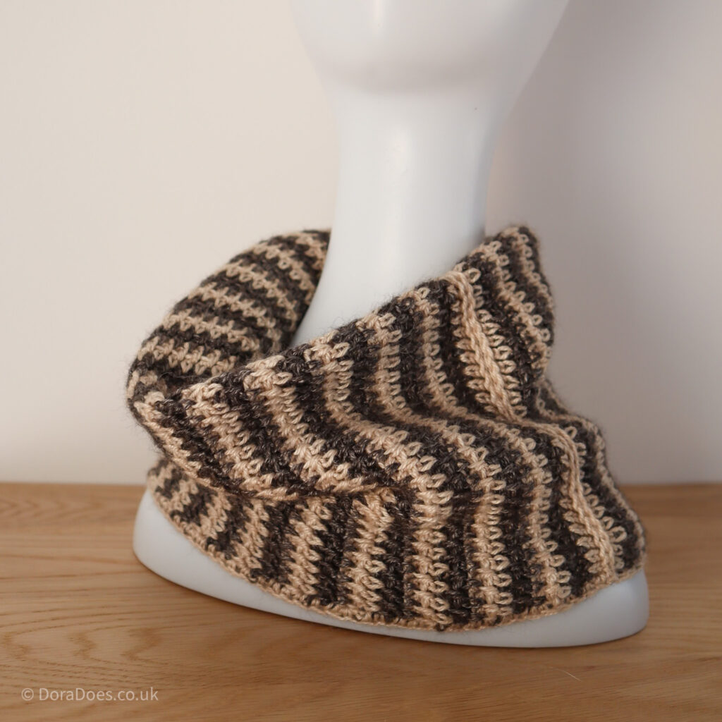 A crochet cowl is doubled over and hung around the neck of a mannequin. It gas jagged vertical stripes where they colours overlap and an intentional seam where the two sides are joined.