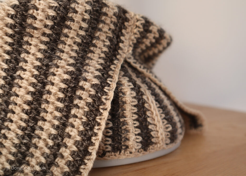 A close up of the crochet stitch pattern used in the between the thorns crochet cowl. You can see dark grey brown yarn contrasted with a champagne cream colour which highlights how the stitches are crocheted between the posts.
