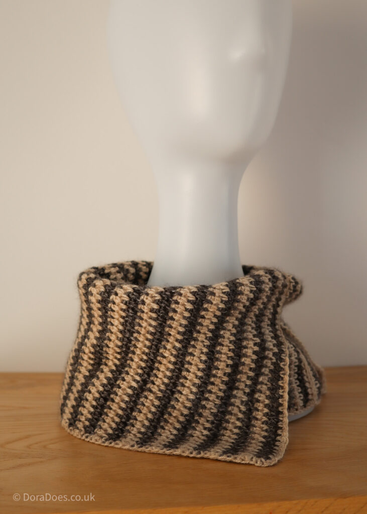 A crochet cowl with a split along one hem is worn folded over with the vertical stripes worn on a slight diagonal. The crochet stitches, made with charcoal and cream yarn interlock to create a kind of spiked zig zag stripe.