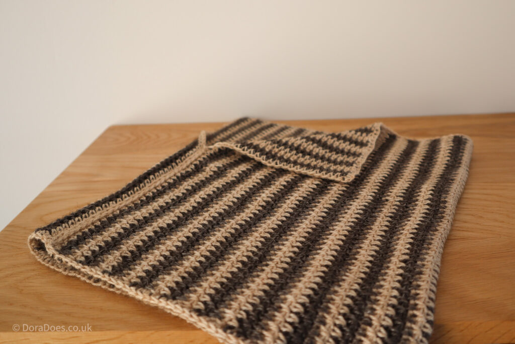 A sample of the ‘between the thorns’ crochet cowl lies flat on a wooden surface with one corner turned down. The cowl has vertical jagged stripes where the crochet stitches are made between the posts, in cream and charcoal brown contrasting yarns.