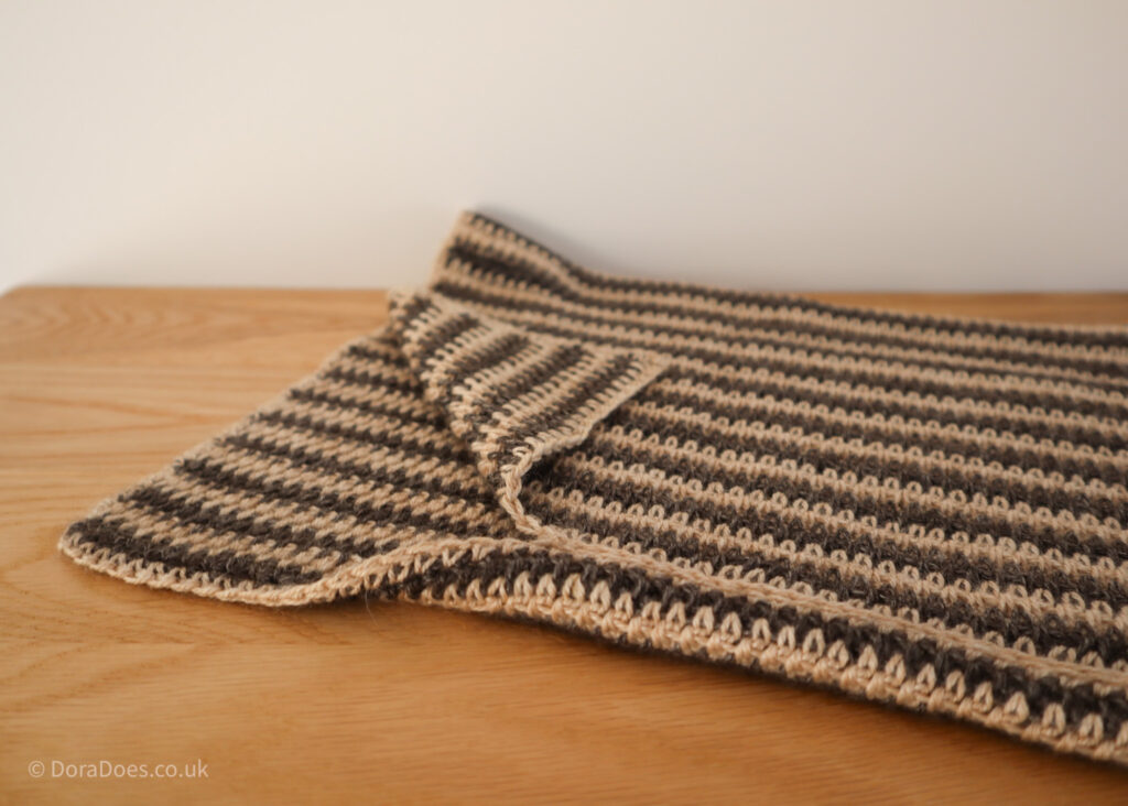 A crochet cowl is laid flat on a wooden surface with the corner of the collar folded over. The neck-warmer is made with two colour of yarn to make a vertical stripe pattern.