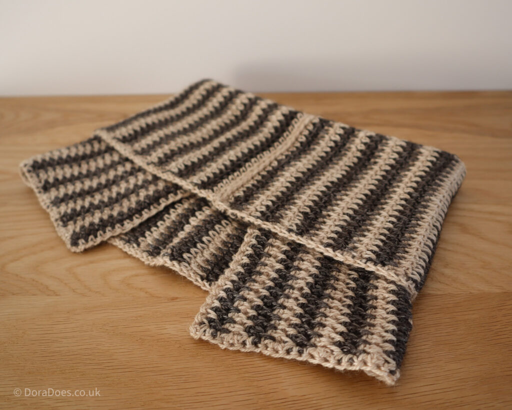 A crochet cowl made with vertical stripes is laid on a flat wooden surface. The top of the cowl is folded over with the bottom split collar poking out looking almost like a bow. The stripes are made with contrasting cream and dark brown yarn.