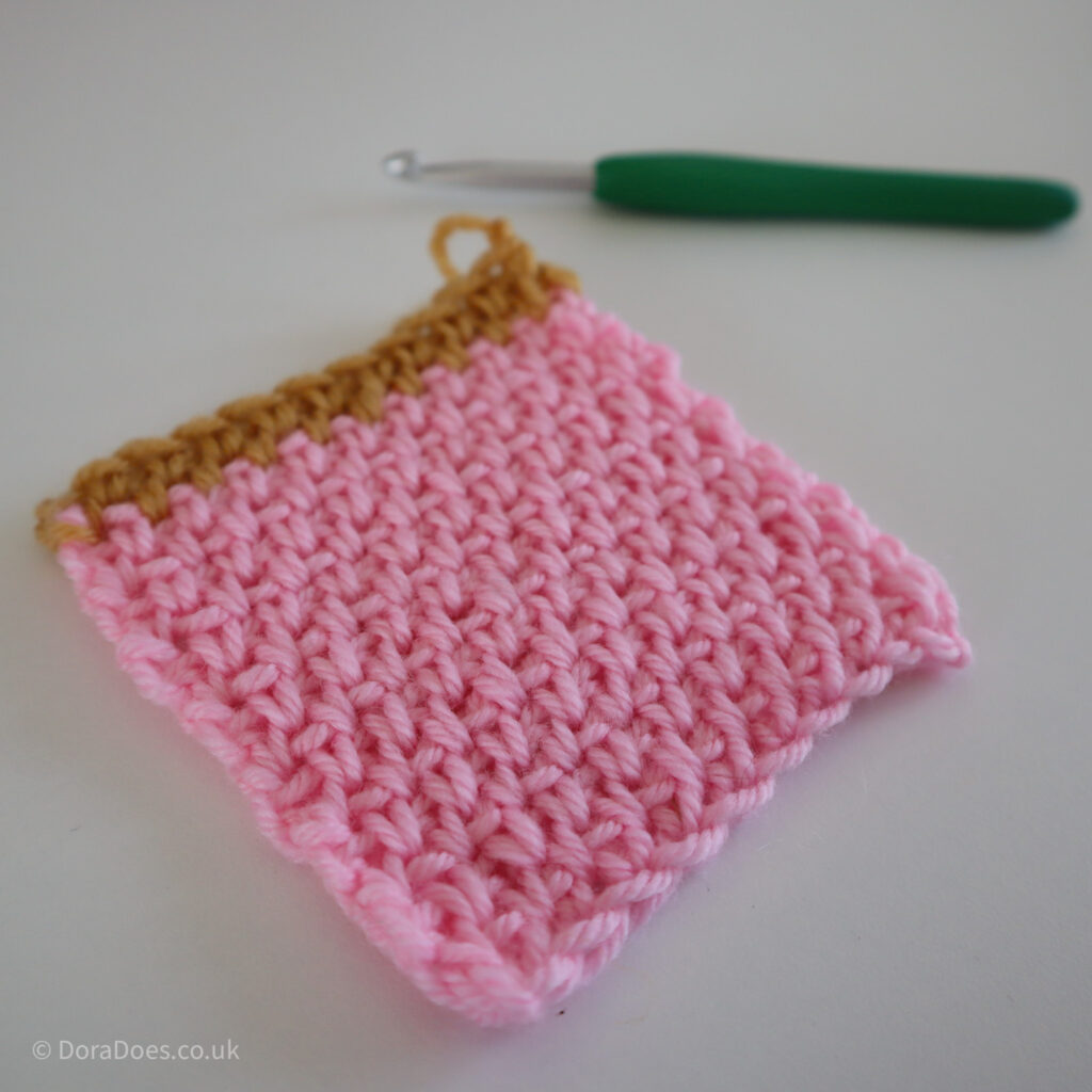A square pink crochet swatch of single crochets between the posts with a mustard yellow stripe of two rows.