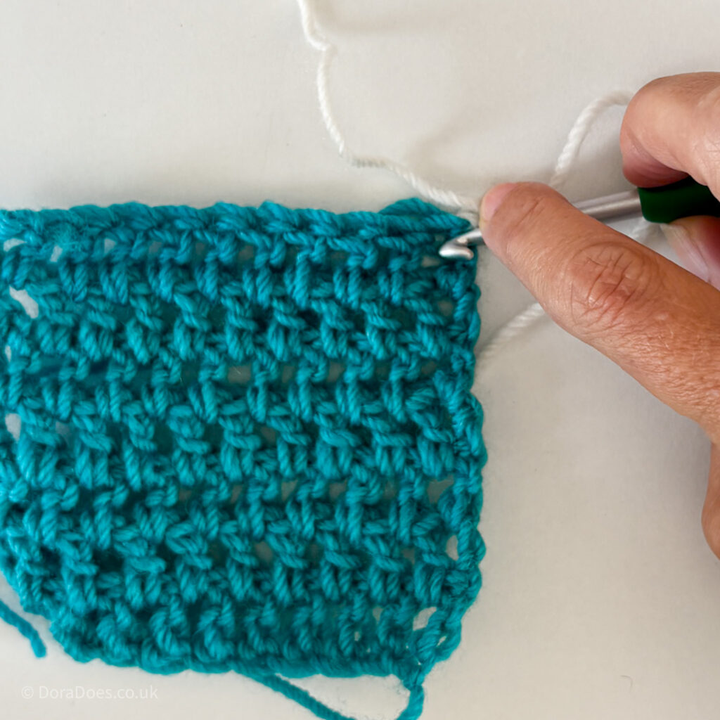 A turquoise swatch of double crochets worked between the posts with a crochet hook pointing to the spot where it should be inserted between the posts.