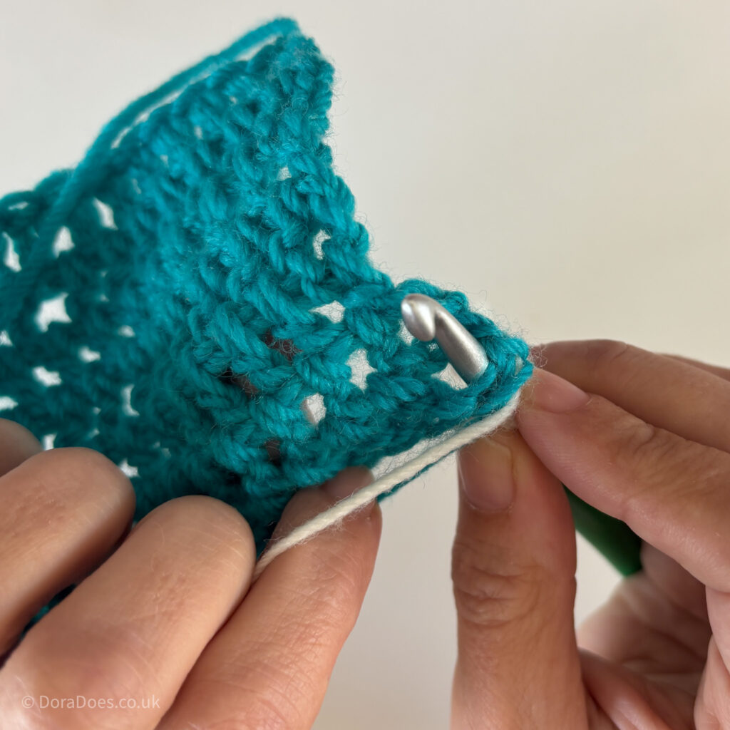A crochet hook inserted through a tortoise swatch of double crochets showing the reverse side of the swatch showing where the hook should come through.