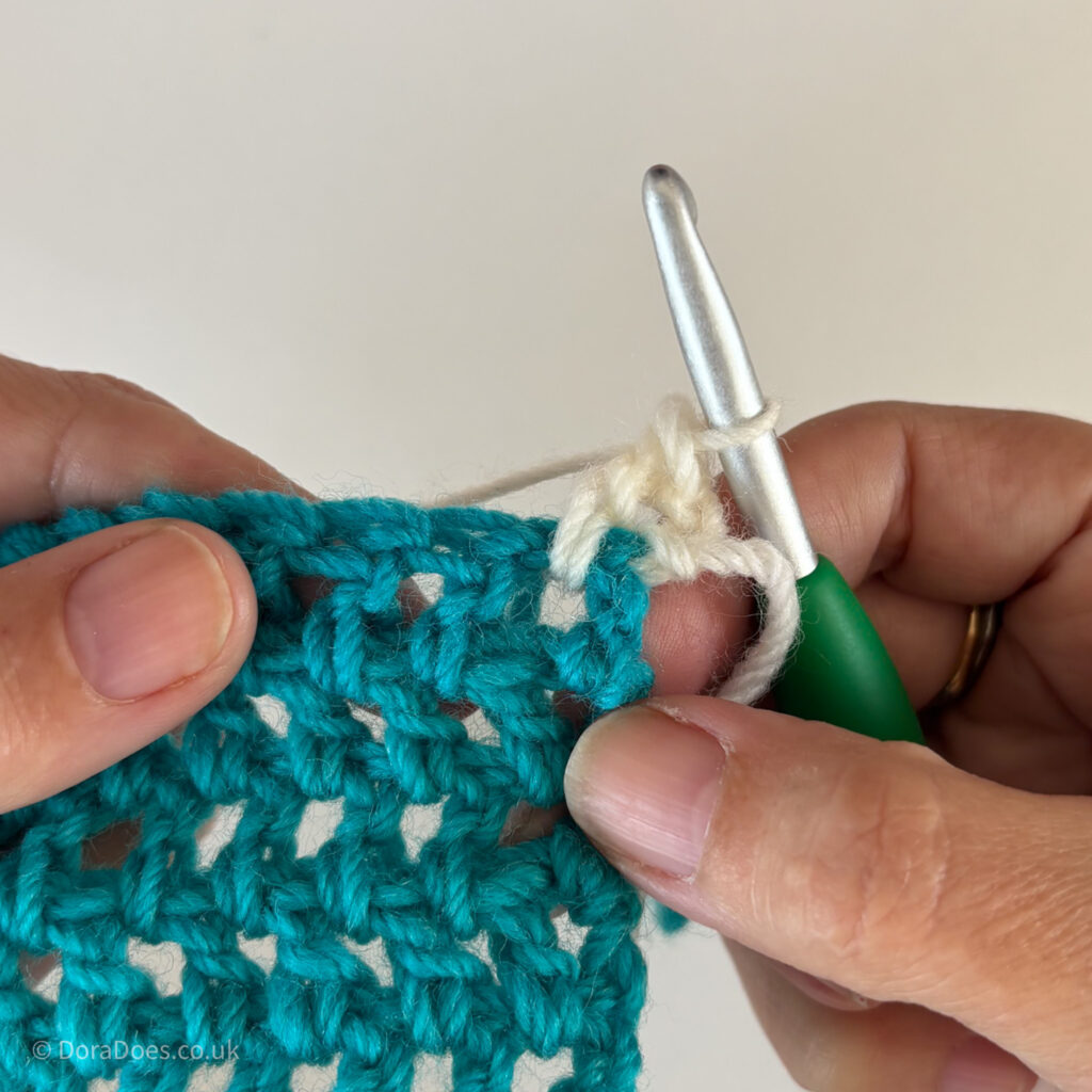 A close up of a double crochet stitch made in cream yarn between the posts of double crochets made in teal yarn.