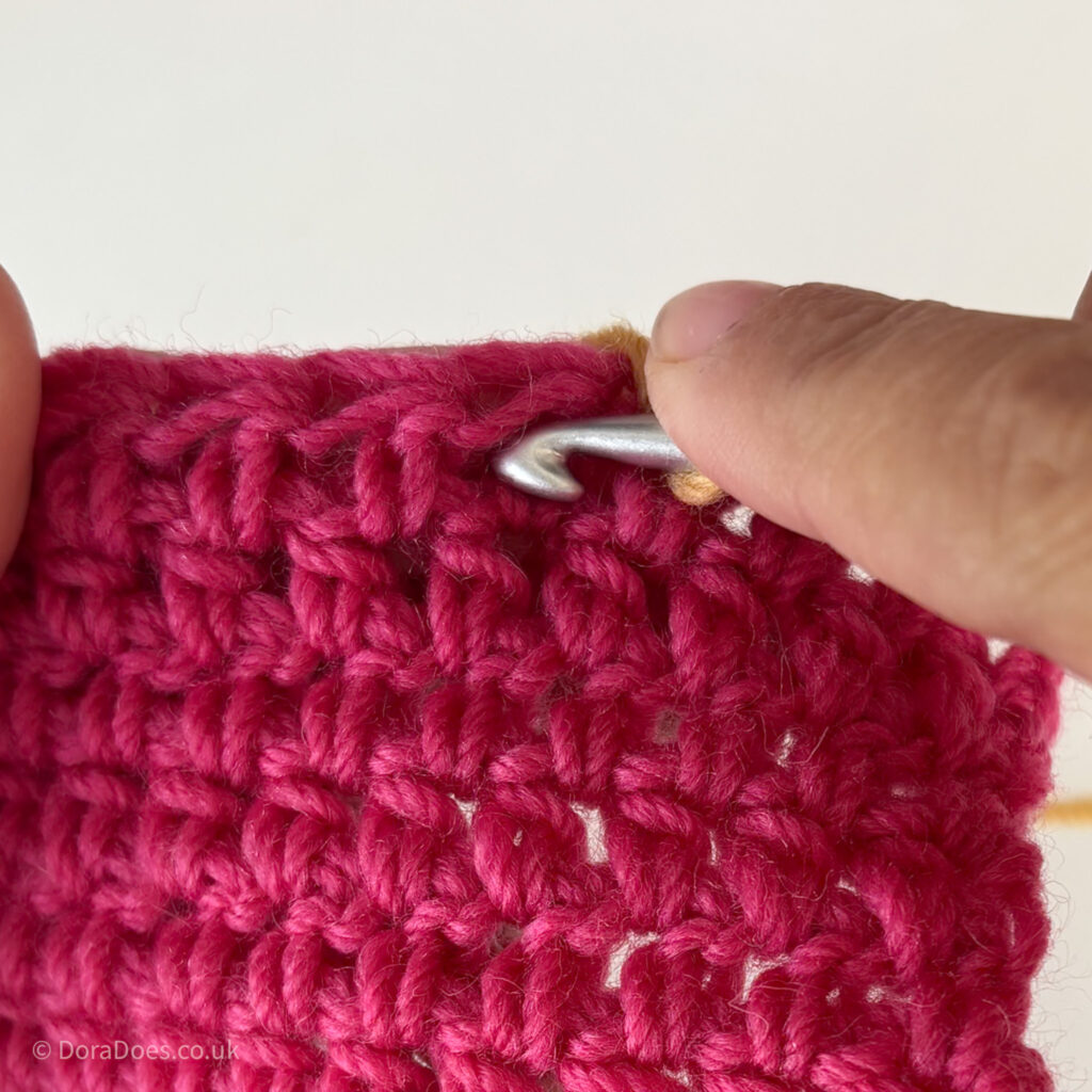 A close up showing where to insert the crochet hook when working between the posts. A hook points so it spot in a pink swatch.