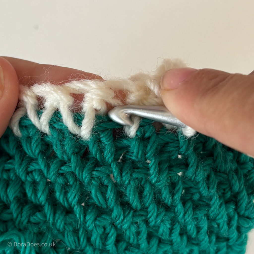 The tip of a crochet hook the pointed at the space where it should be inserted to work between the stitches of a green extended singe crochet swatch.